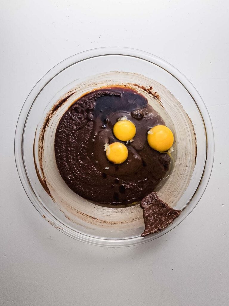 Glass bowl with brownie batter and three raw eggs on top, ready to be mixed for delicious fudgy brownie cookies, on a white surface.
