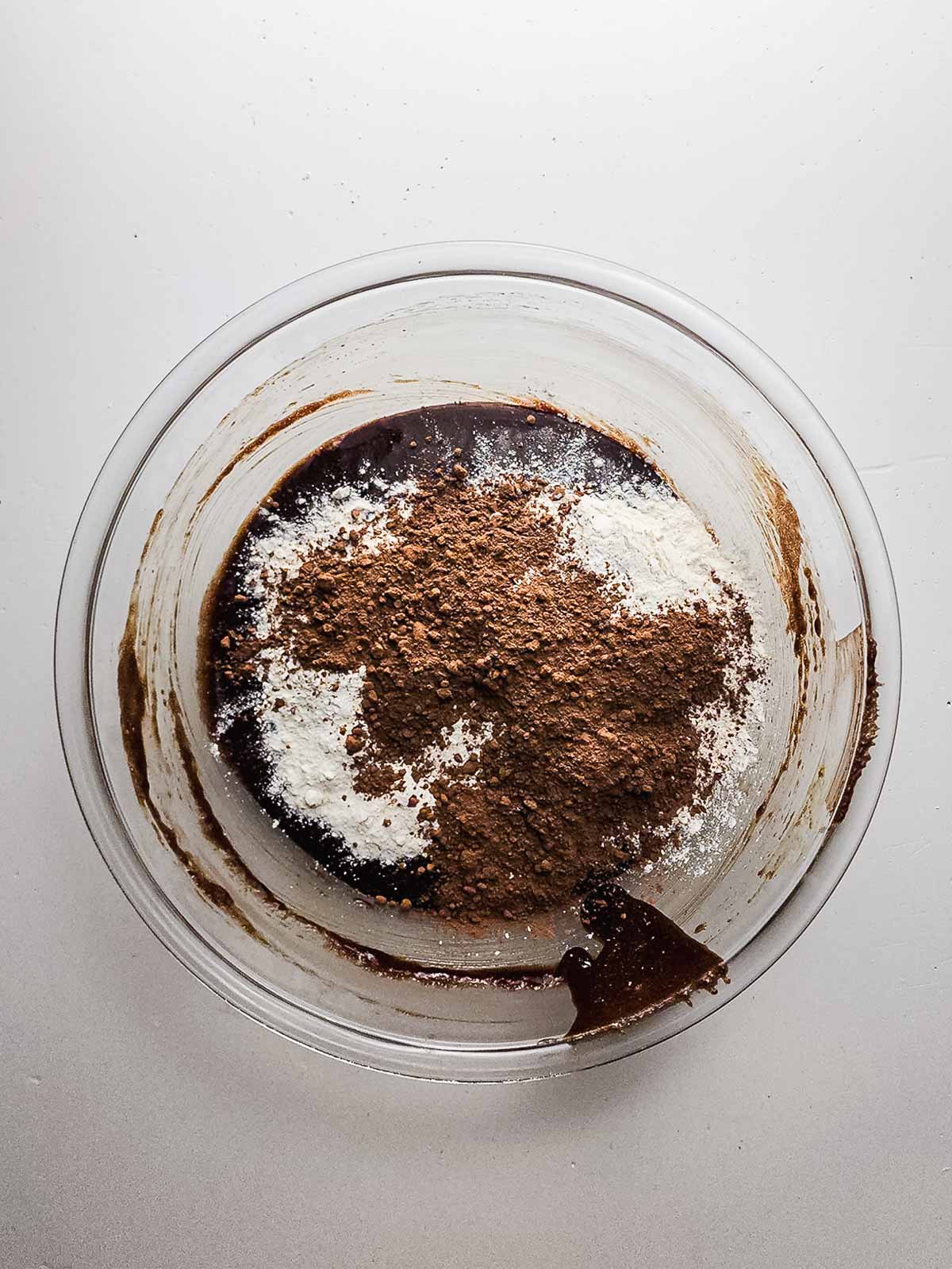 A glass bowl with brownie batter, cocoa powder, and flour on top, ready to be mixed for delicious Fudgy Brownie Cookies.