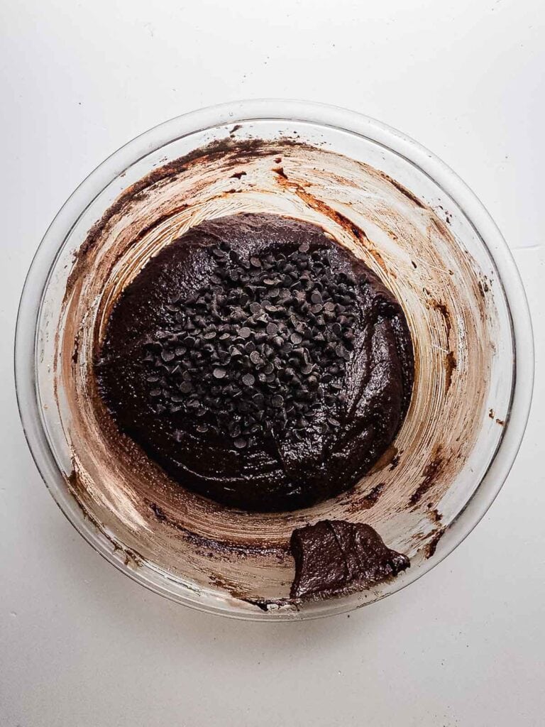 A glass bowl with chocolate brownie batter topped with chocolate chips on a white surface, perfect for making rich and fudgy brownie cookies.