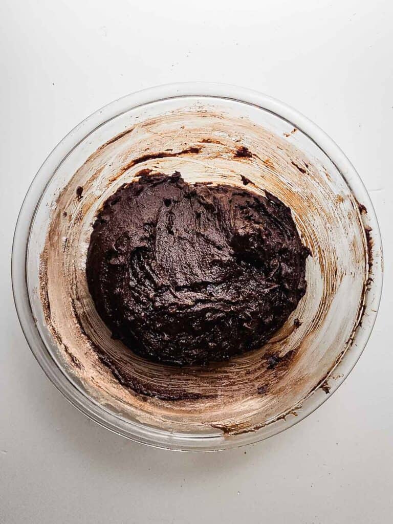 A glass bowl with chocolate brownie batter, perfect for making fudgy brownie cookies, partially mixed and sitting on a white surface.