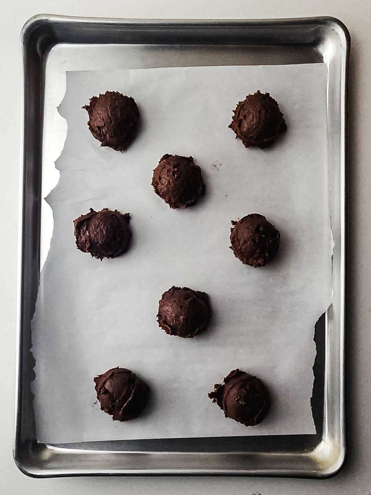 Eight scoops of fudgy brownie cookie dough on parchment paper rest on a baking sheet, ready to be baked into irresistible treats.