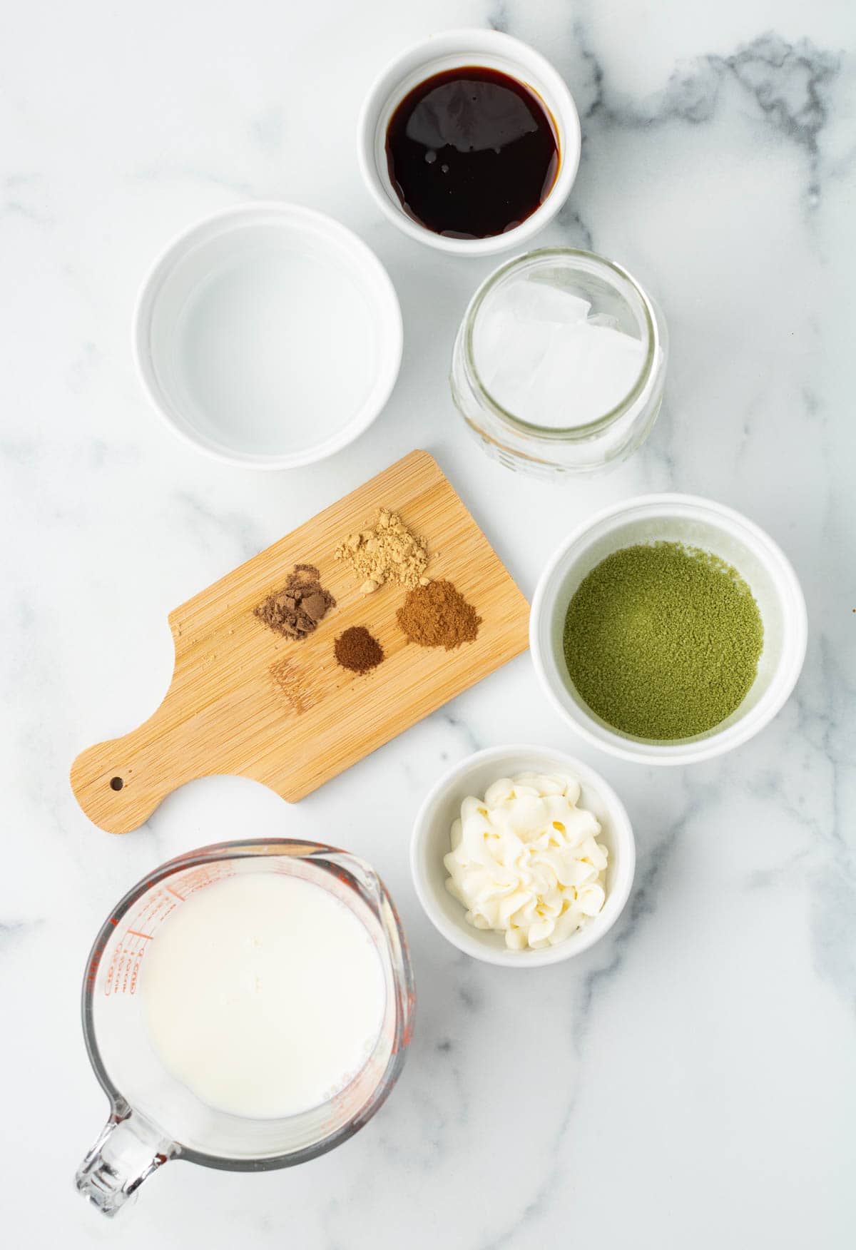 Various ingredients including spices, Gingerbread Matcha powder, milk, and syrups are artfully arranged on a white marble surface.
