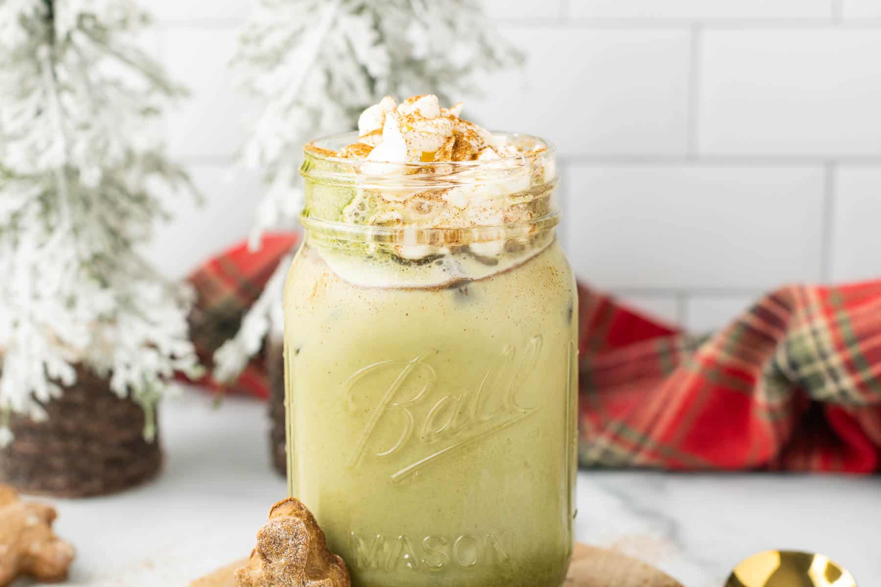 Iced Gingerbread Matcha Latte in a mason jar topped with whipped cream and cinnamon, with festive decor in the background.