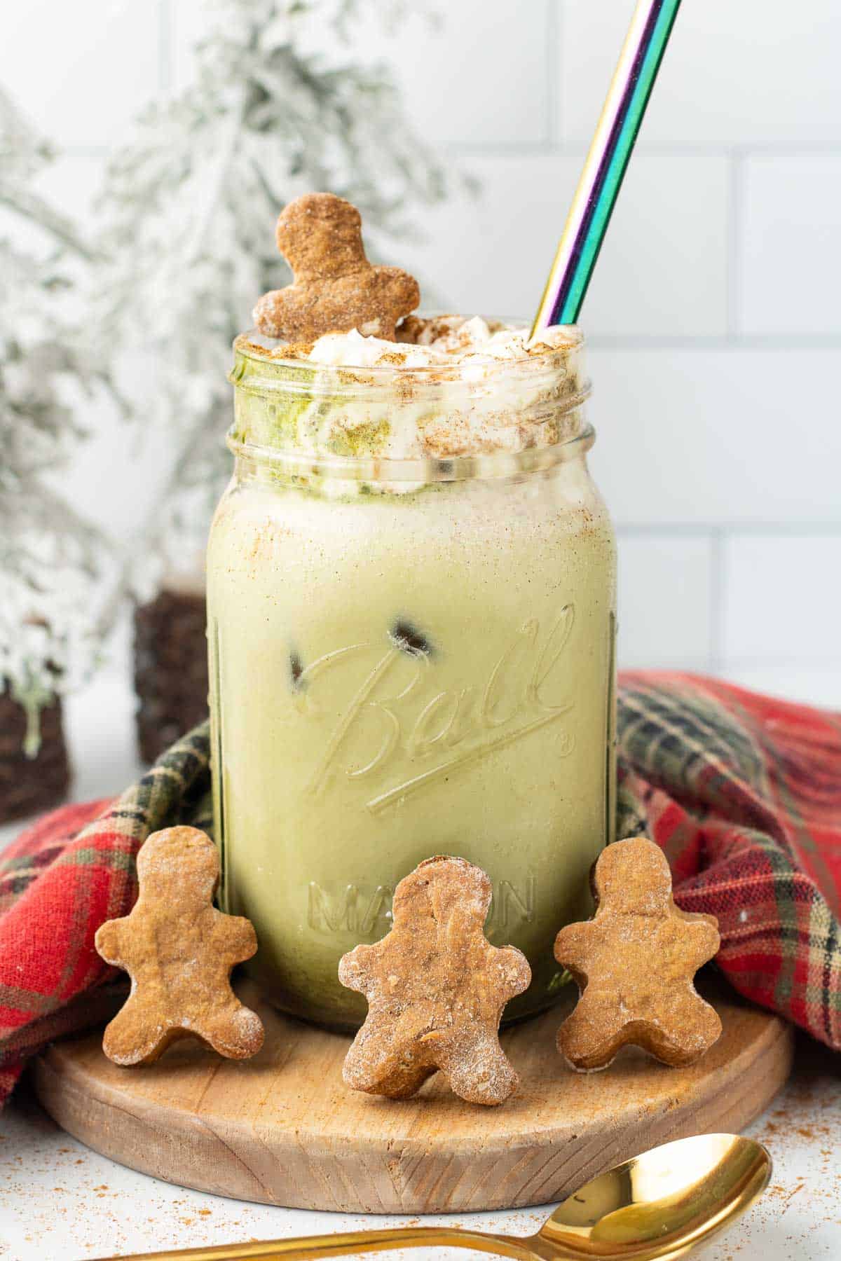 Iced Gingerbread Matcha Latte in a mason jar with whipped cream, topped with mini gingerbread cookies and a striped straw.