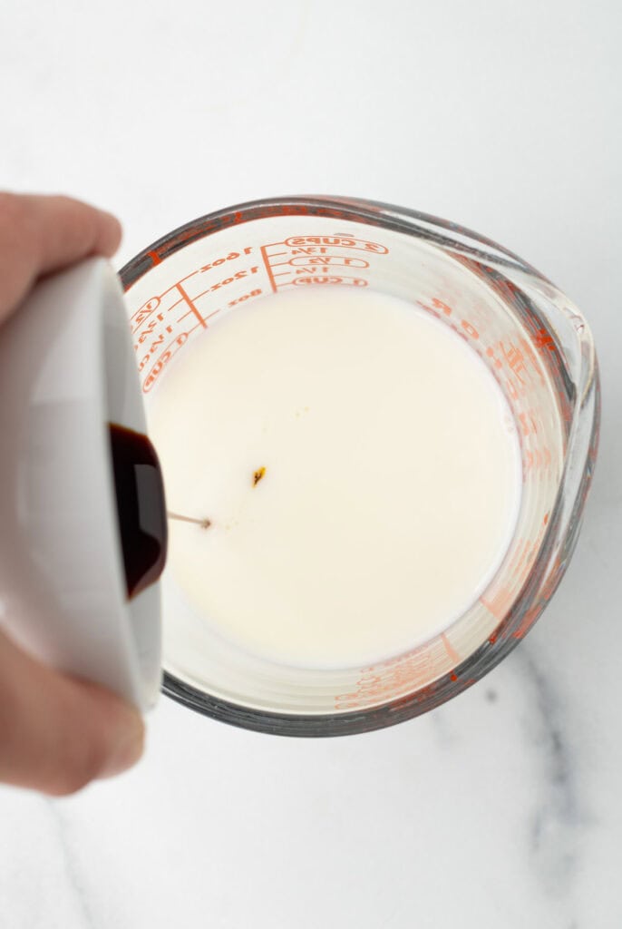 A hand pours vanilla extract into a glass measuring cup filled with milk on a white surface, preparing the base for a cozy gingerbread treat.
