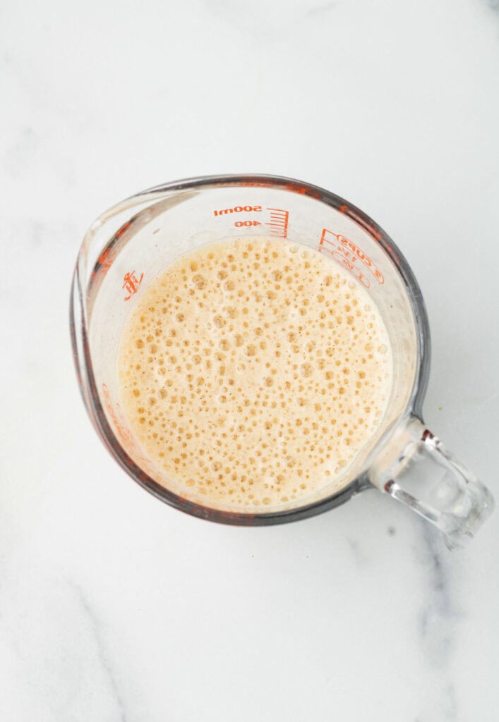 A glass measuring cup filled with a bubbly, light brown Gingerbread Matcha Latte sits on a white surface.