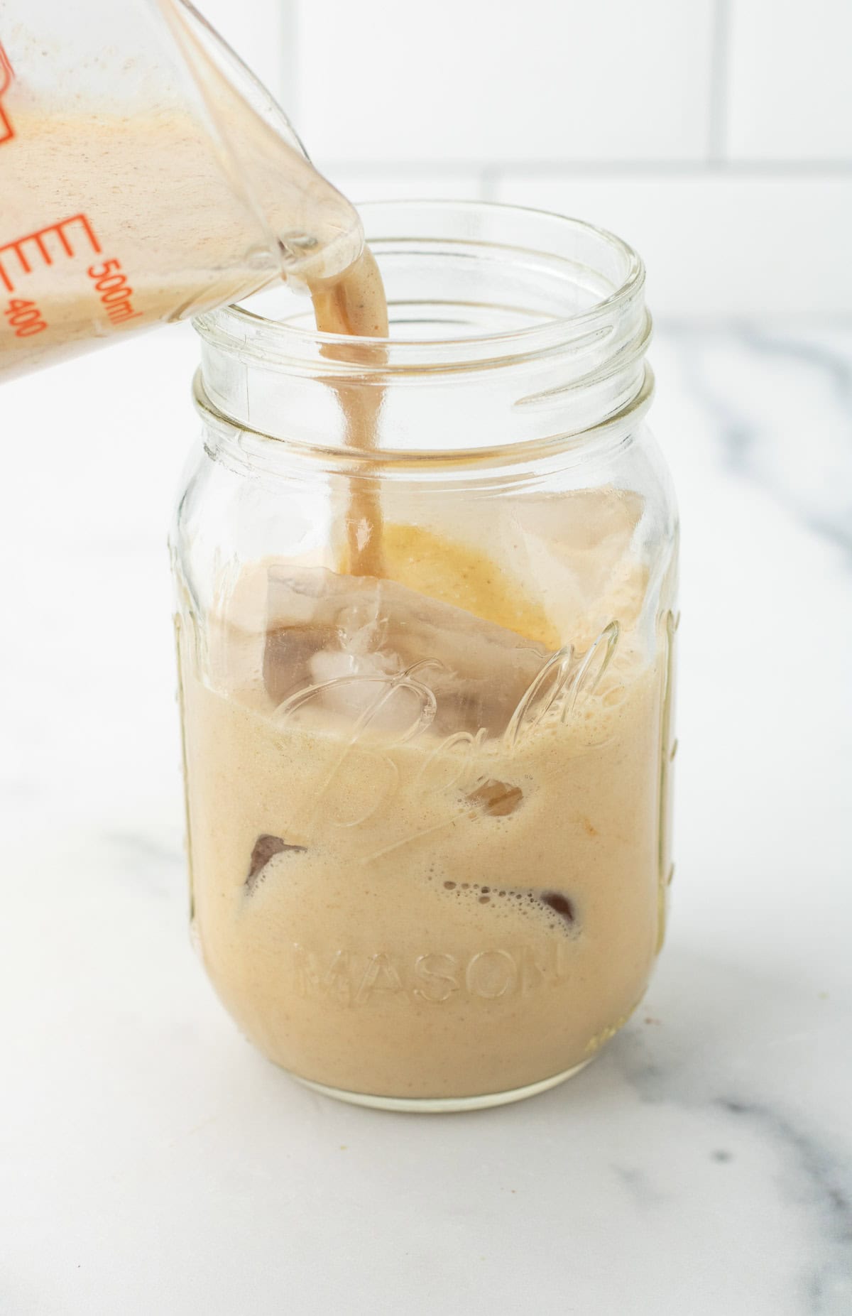 Creamy iced coffee being poured from a measuring cup into a mason jar with ice cubes on a marble counter, perfect for pairing with your favorite matcha latte or gingerbread treat.