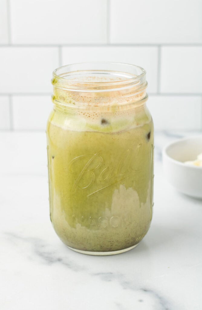 A mason jar filled with iced Gingerbread Matcha Latte sits on a white marble counter, set against a white tiled background.