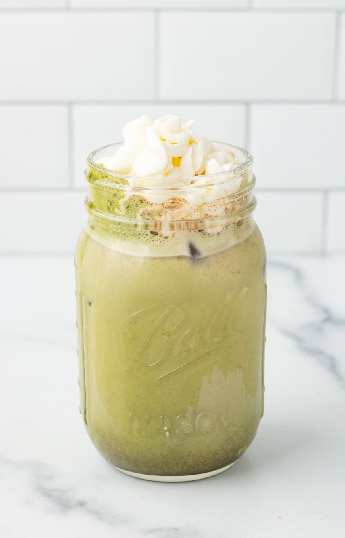 Iced Gingerbread Matcha Latte in a mason jar topped with whipped cream, on a marble counter with a tiled background.