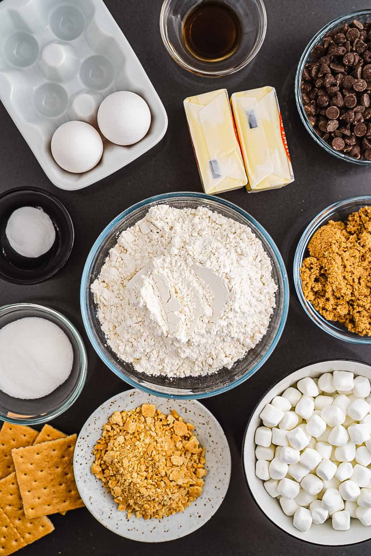 Baking ingredients on a dark surface: flour, eggs, butter, chocolate chips, sugars, marshmallows, and graham crackers&mdash;everything you need for the perfect S'mores cookies recipe.