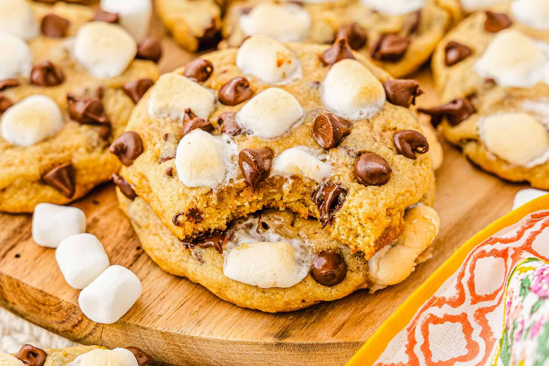 Chocolate chip and marshmallow S'mores cookies on a wooden board, with one cookie showing a bite taken out&mdash;a tempting twist on the classic s'mores recipe.