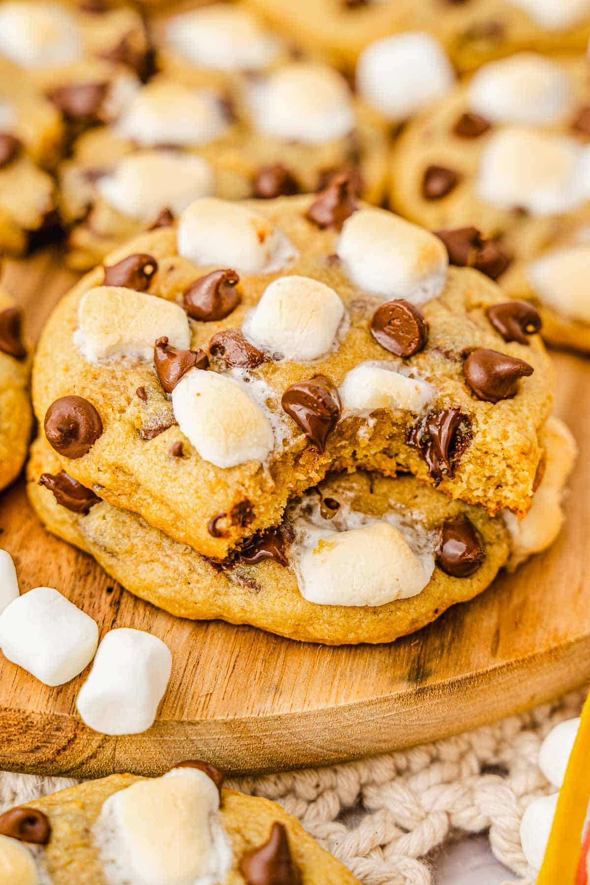 A S'mores Cookies recipe brought to life&mdash;a cookie loaded with chocolate chips and toasted marshmallows, with a bite taken out, resting on a wooden board.