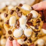 A hand holding a gooey S'mores Cookies recipe creation topped with melted marshmallows and chocolate chips.