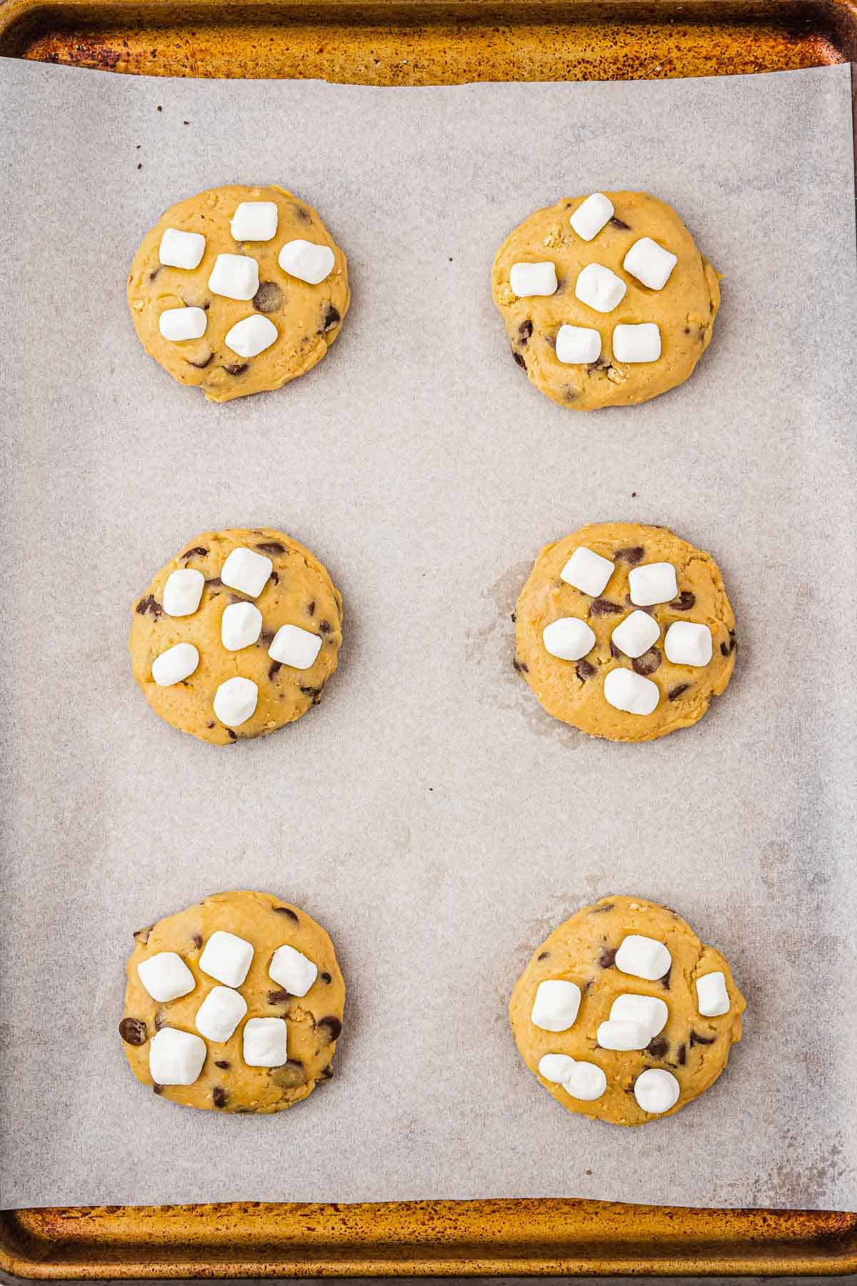 Six unbaked S'mores cookies, filled with chocolate chips and mini marshmallows, rest on a parchment-lined baking sheet&mdash;perfect for trying out your next s'mores recipe.