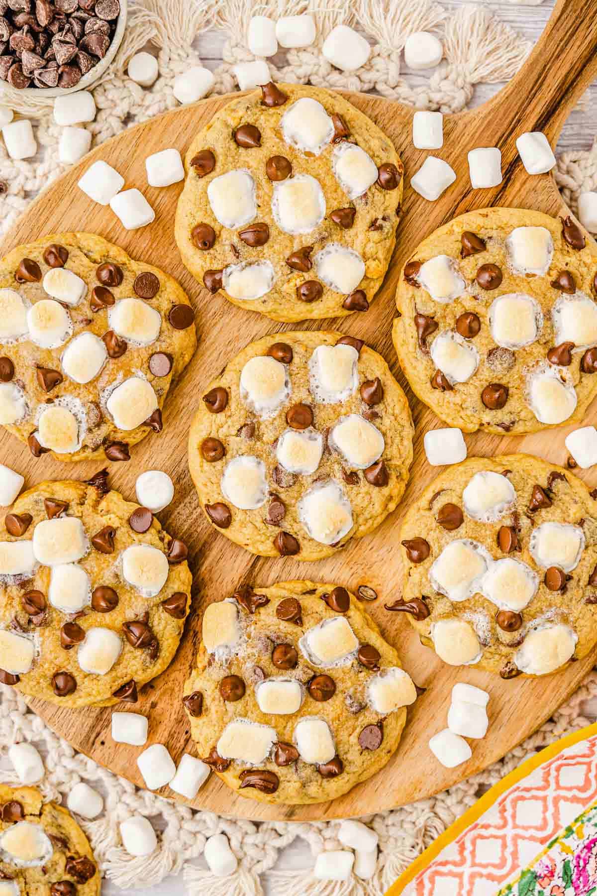 Cookies topped with chocolate chips and mini marshmallows arranged on a wooden board, evoking a classic S'mores Cookies recipe, with extra marshmallows scattered around.