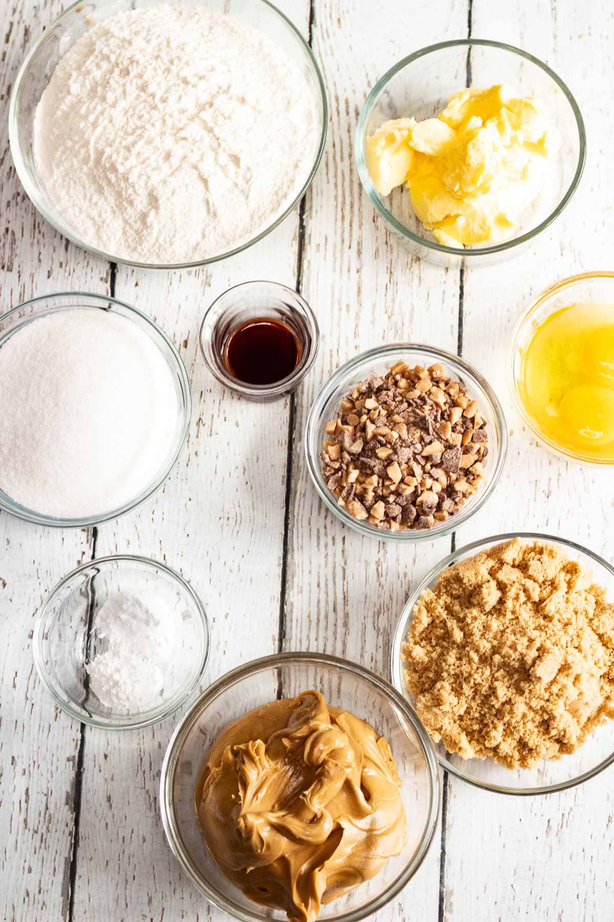 Bowls of flour, butter, sugar, brown sugar, vanilla, toffee bits, baking powder, egg, and peanut butter on wood&mdash;perfect for making delicious Peanut Butter Cookies.