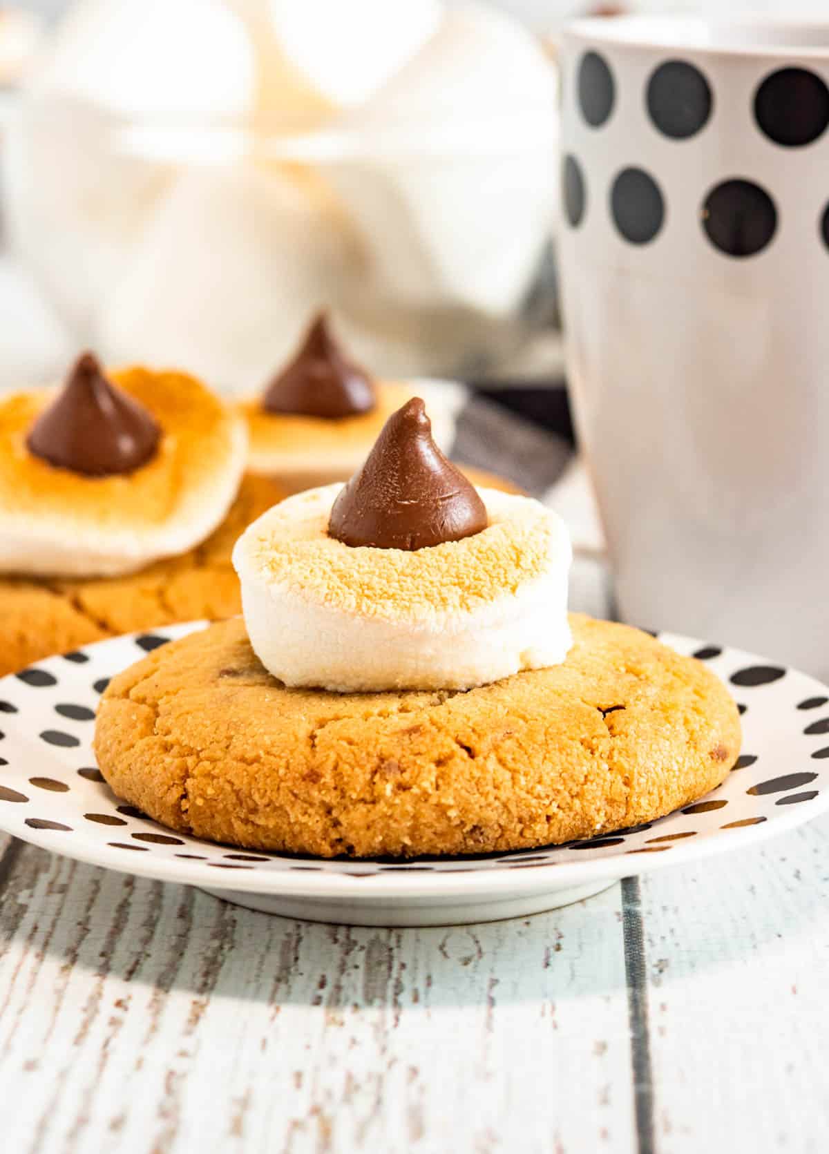 A peanut butter cookie topped with a toasted marshmallow and a chocolate candy sits on a white plate with black dots.