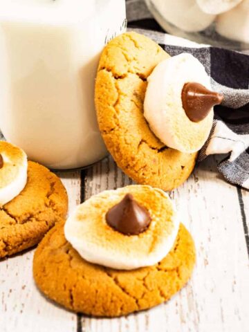Three Blossom Cookies with toasted marshmallows and chocolate sit beside a glass of milk with straws and a bowl of marshmallows.