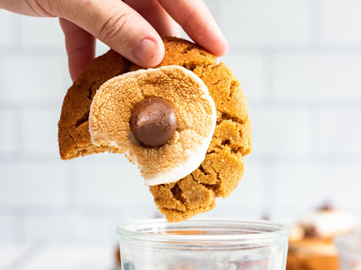 A hand dips a s'mores cookie with a bite missing into a glass of milk, bringing to mind the comforting taste of Peanut Butter Cookies.