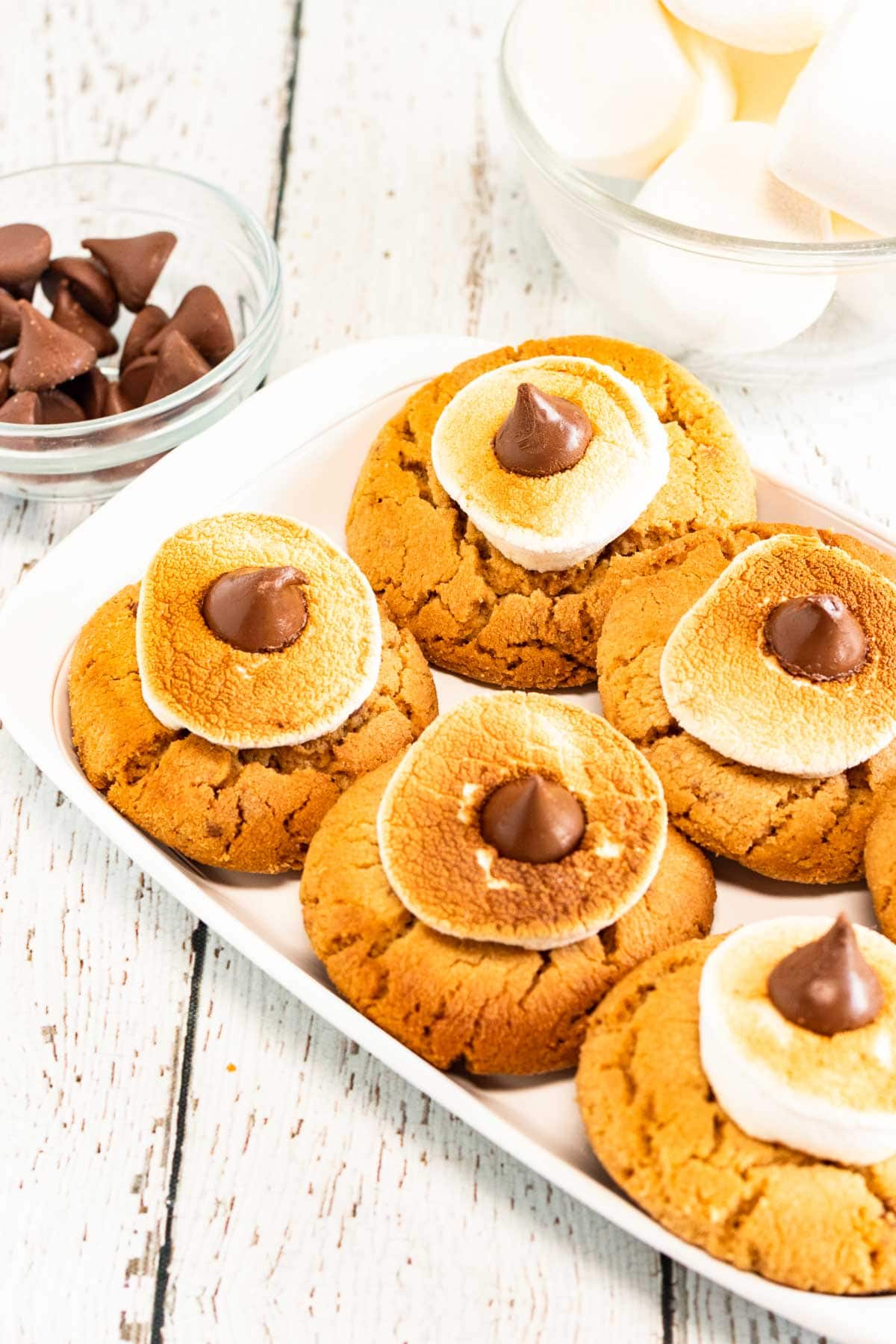 Plate of Blossom Cookies topped with toasted marshmallows and chocolate kisses, next to bowls of chocolate and marshmallows&mdash;perfect for peanut butter cookie lovers.