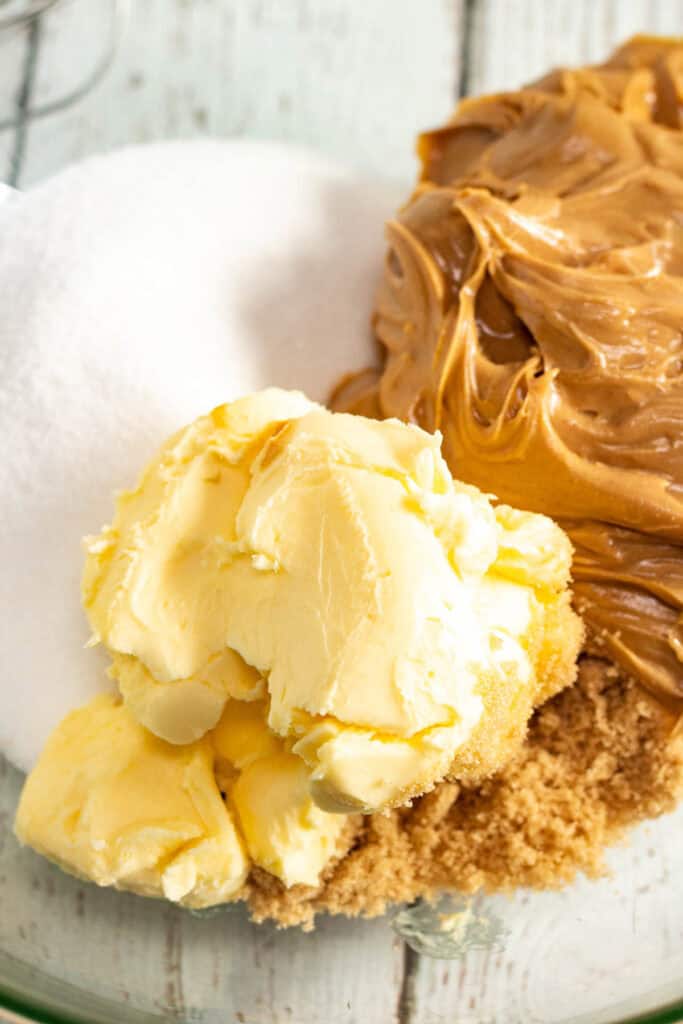 Butter, brown sugar, white sugar, and creamy peanut butter are ready in a glass bowl on a white wooden surface&mdash;perfect for making classic Peanut Butter Cookies or festive Blossom Cookies.