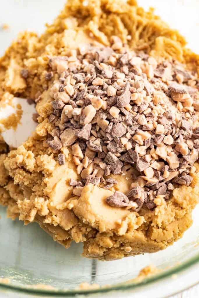 Cookie dough with chopped toffee bits and chocolate pieces on top in a glass mixing bowl, perfect for creating irresistible peanut butter cookies.