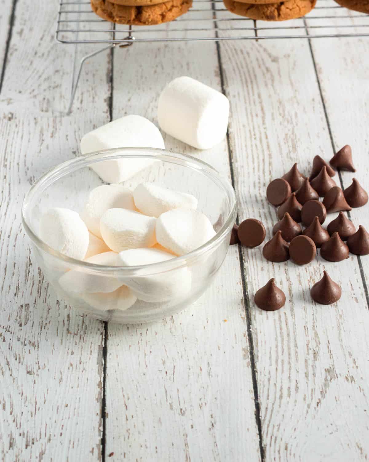 A glass bowl of marshmallows next to chocolate chips on a white wooden surface, with peanut butter cookies and other treats in the background.