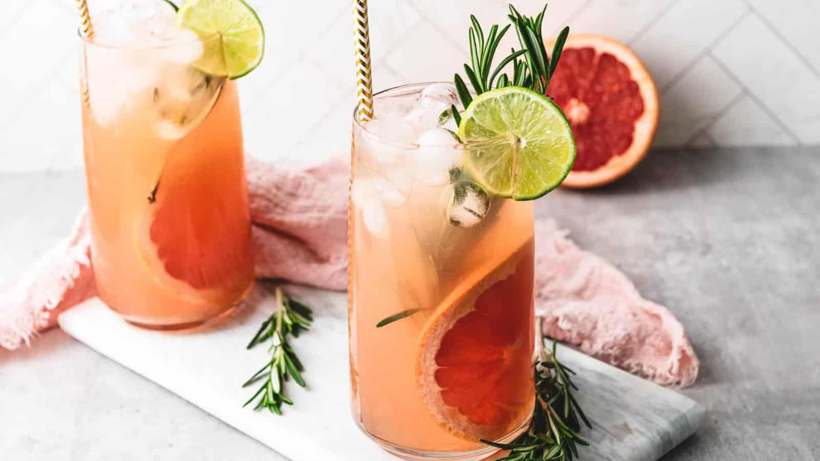 Two grapefruit cocktails with ice, lime slices, and rosemary garnish on a marble board, with a grapefruit in back.