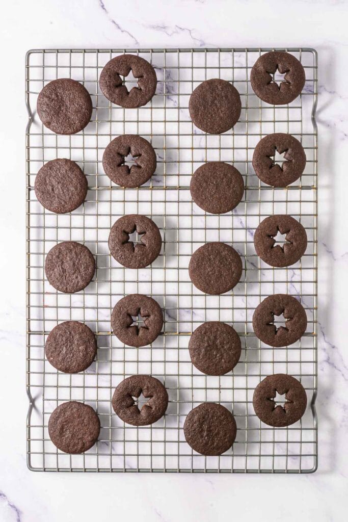 Chocolate Linzer Cookies, some with star-shaped cutouts, rest on a cooling rack over a white marble surface.