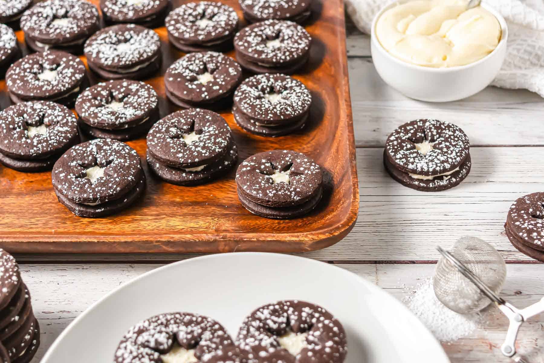 Chocolate Linzer Cookies with powdered sugar on top, filled with cream, are beautifully arranged on a wooden tray and white plate. Try this delicious Linzer Cookies recipe for a delightful treat.