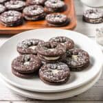 Chocolate Linzer Cookies with cream filling, dusted with powdered sugar, are stacked on white plates.