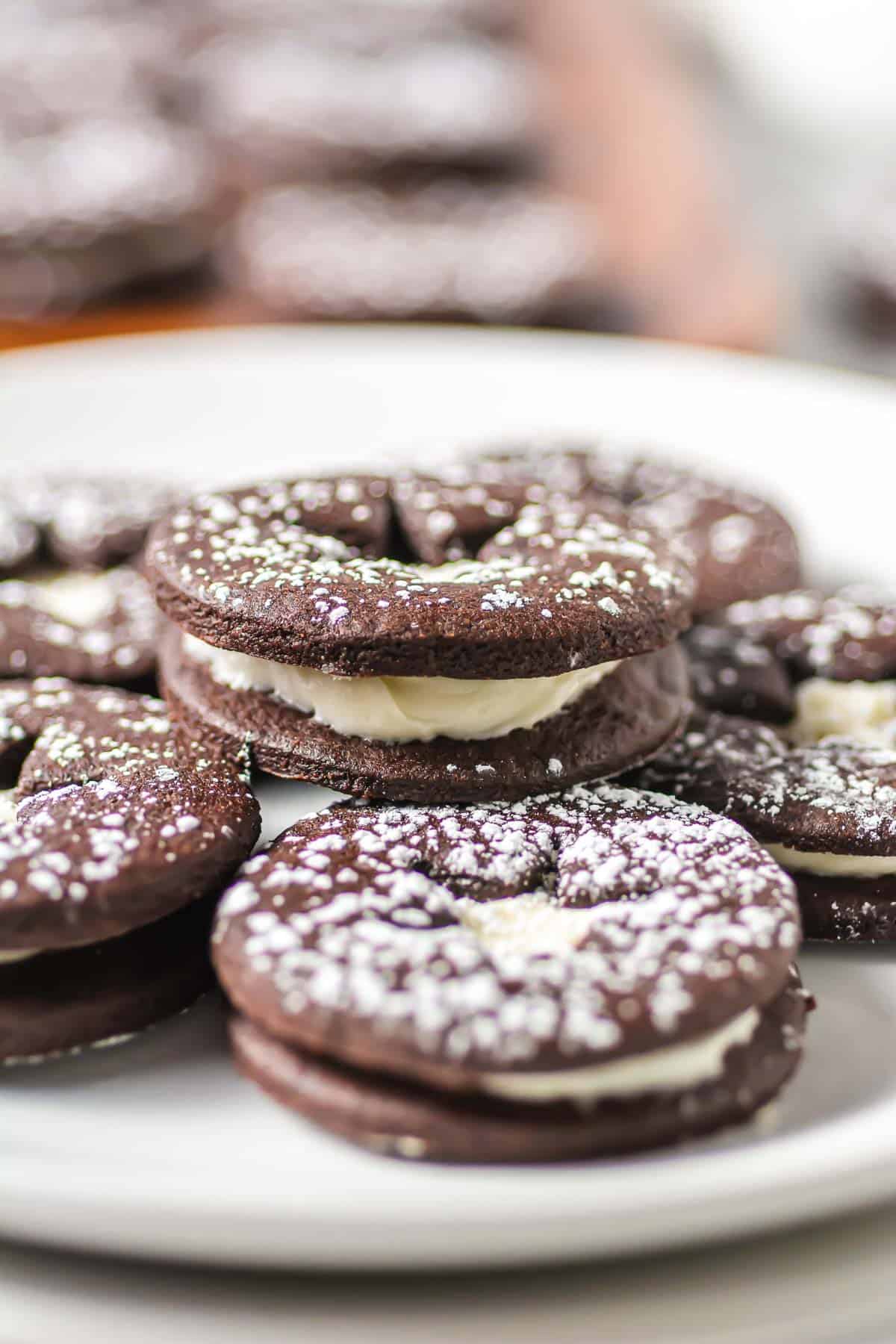 Chocolate Linzer Cookies with white cream filling and powdered sugar on top, stacked neatly on a white plate.