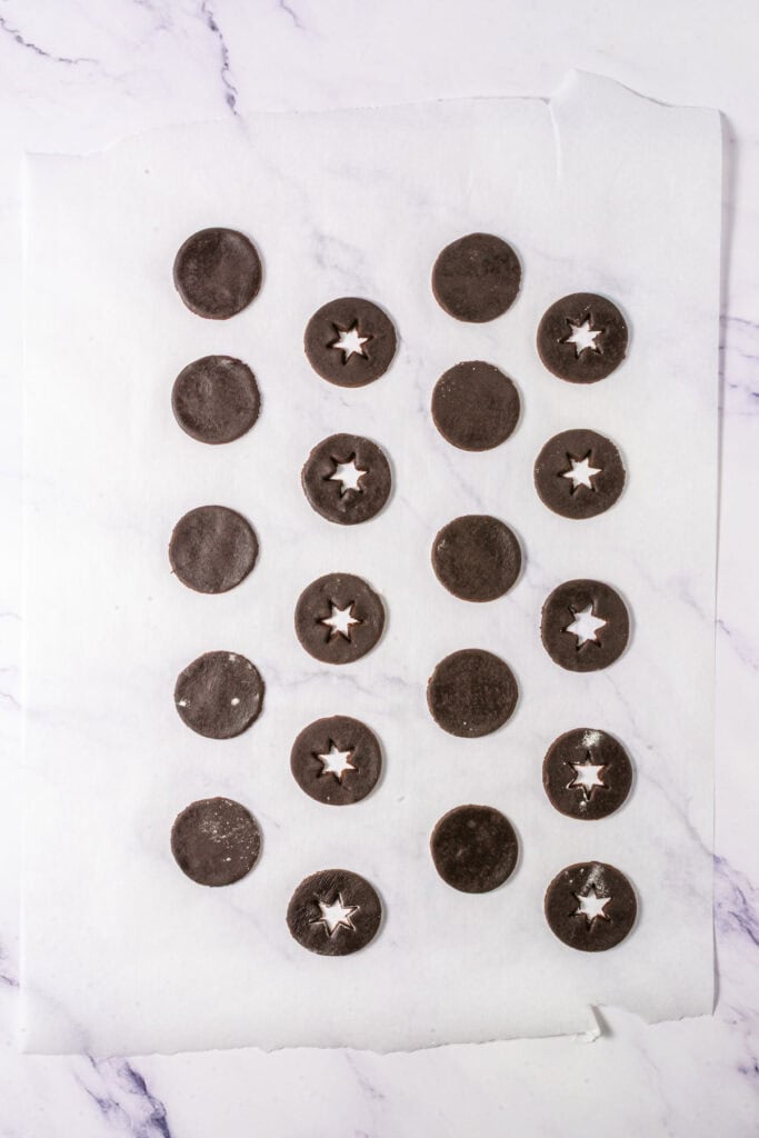 Twenty Chocolate Linzer Cookies, some with star-shaped cutouts, arranged on parchment paper, showcase a delicious twist on the classic Linzer Cookies recipe.