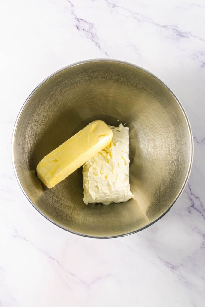 A stick of butter and a block of cream cheese sit in a metal mixing bowl on a marble surface, ready to be transformed into rich Chocolate Linzer Cookies.