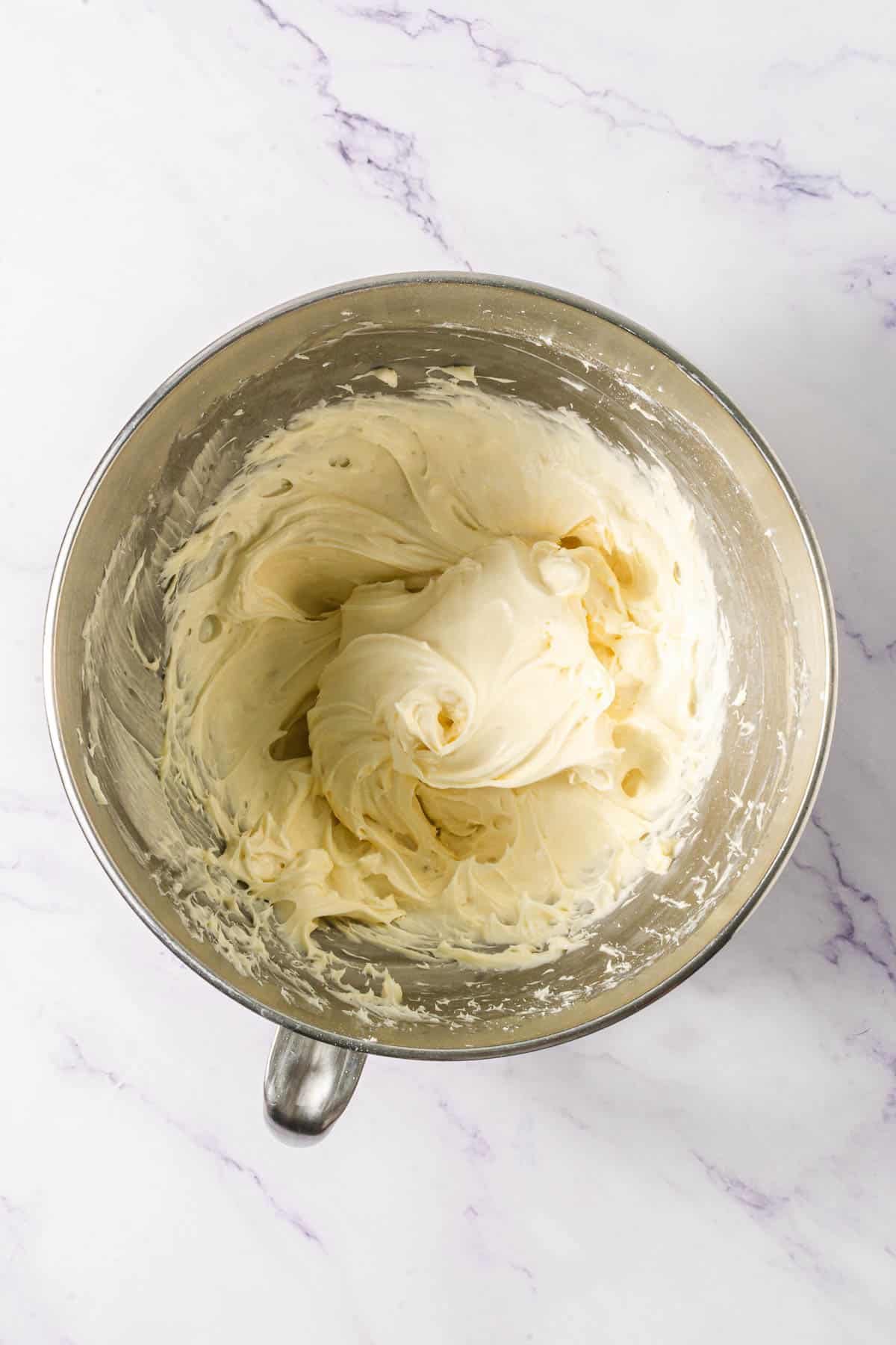 A metal mixing bowl filled with creamy, whipped frosting on a white marble surface, perfect for topping your favorite Chocolate Linzer Cookies.