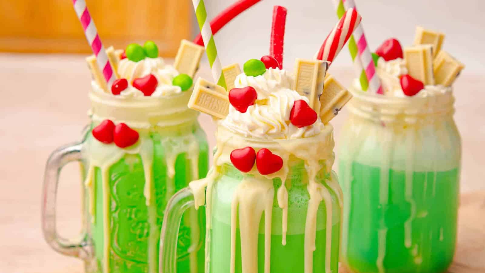 Three green milkshakes in mason jars, topped with whipped cream, candy, and white chocolate pieces.
