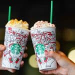 Two hands holding festive Starbucks Holiday Drinks cups with whipped cream toppings, raised in a celebratory toast&mdash;cheering to hot and iced holiday drinks.