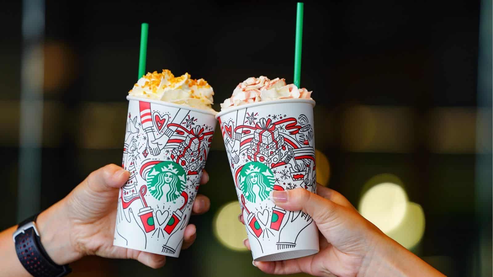 Two hands holding festive Starbucks Holiday Drinks cups with whipped cream toppings, raised in a celebratory toast&mdash;cheering to hot and iced holiday drinks.
