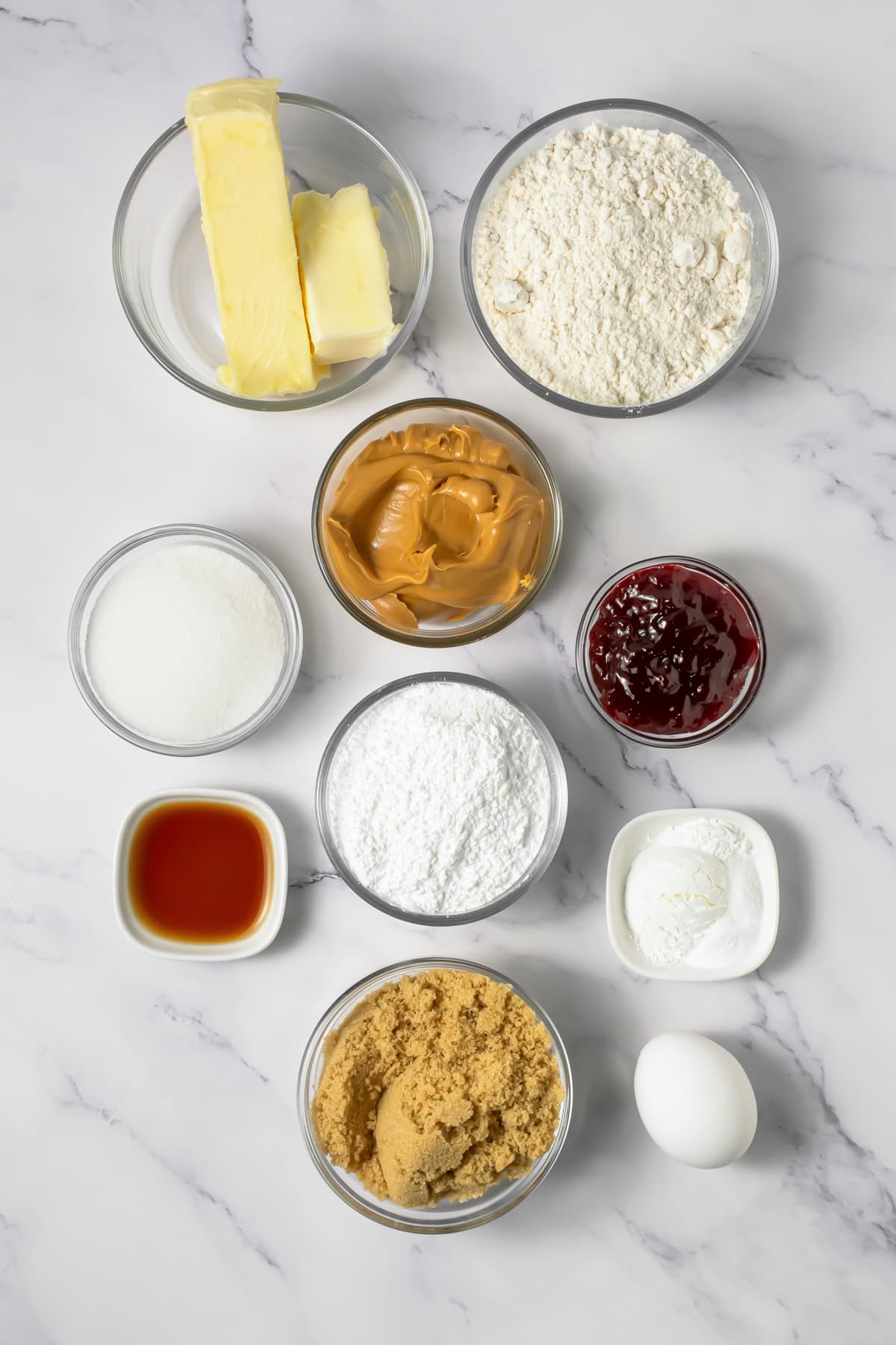 Baking ingredients in bowls: butter, flour, sugar, peanut butter, jam, vanilla, brown sugar, powder, and an egg&mdash;everything you need for Crumbl copycat peanut butter and jelly cookies.