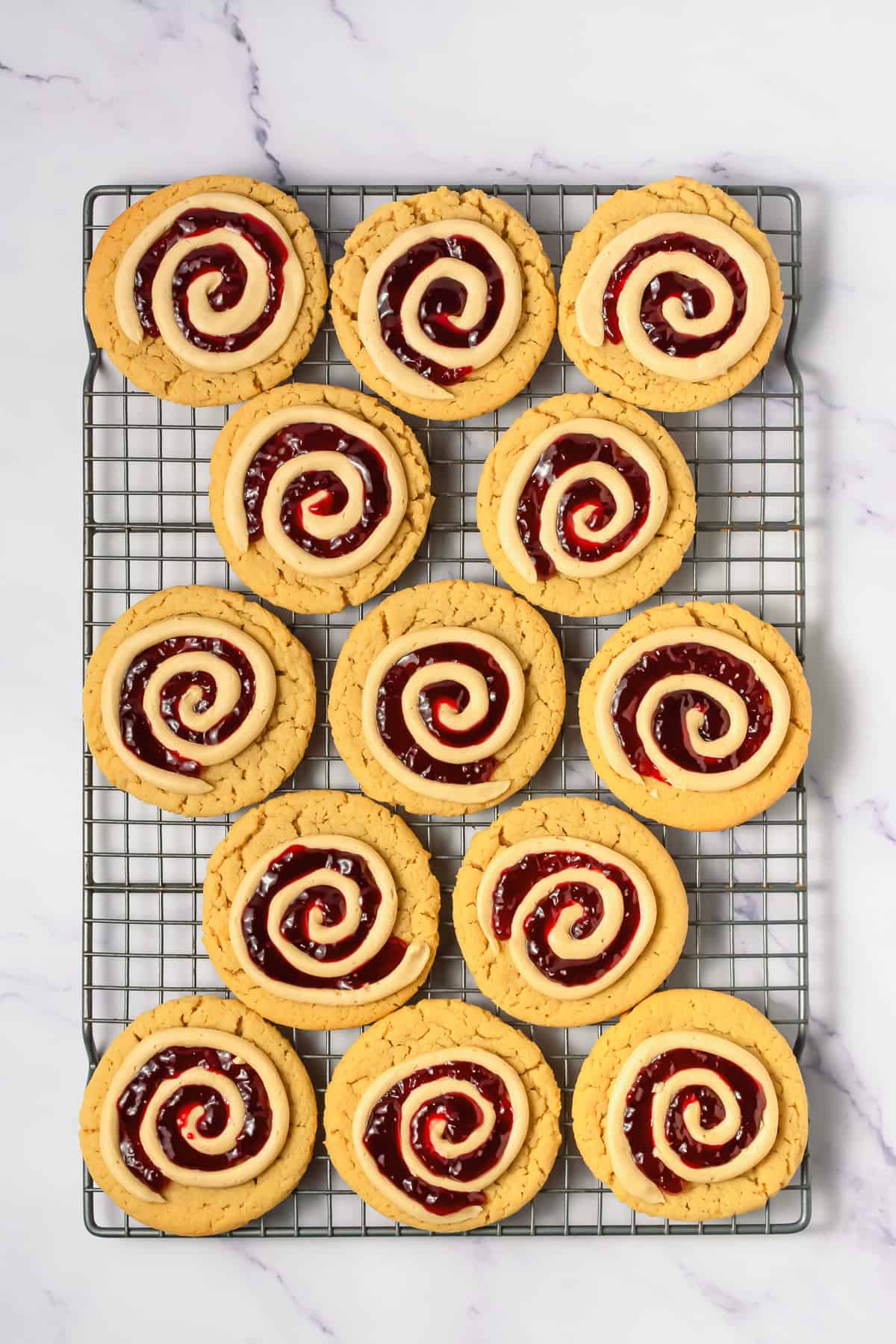 Twelve spiral-iced cookies with red jam, inspired by Crumbl copycat peanut butter and jelly cookies, rest on a cooling rack over a white marble surface.