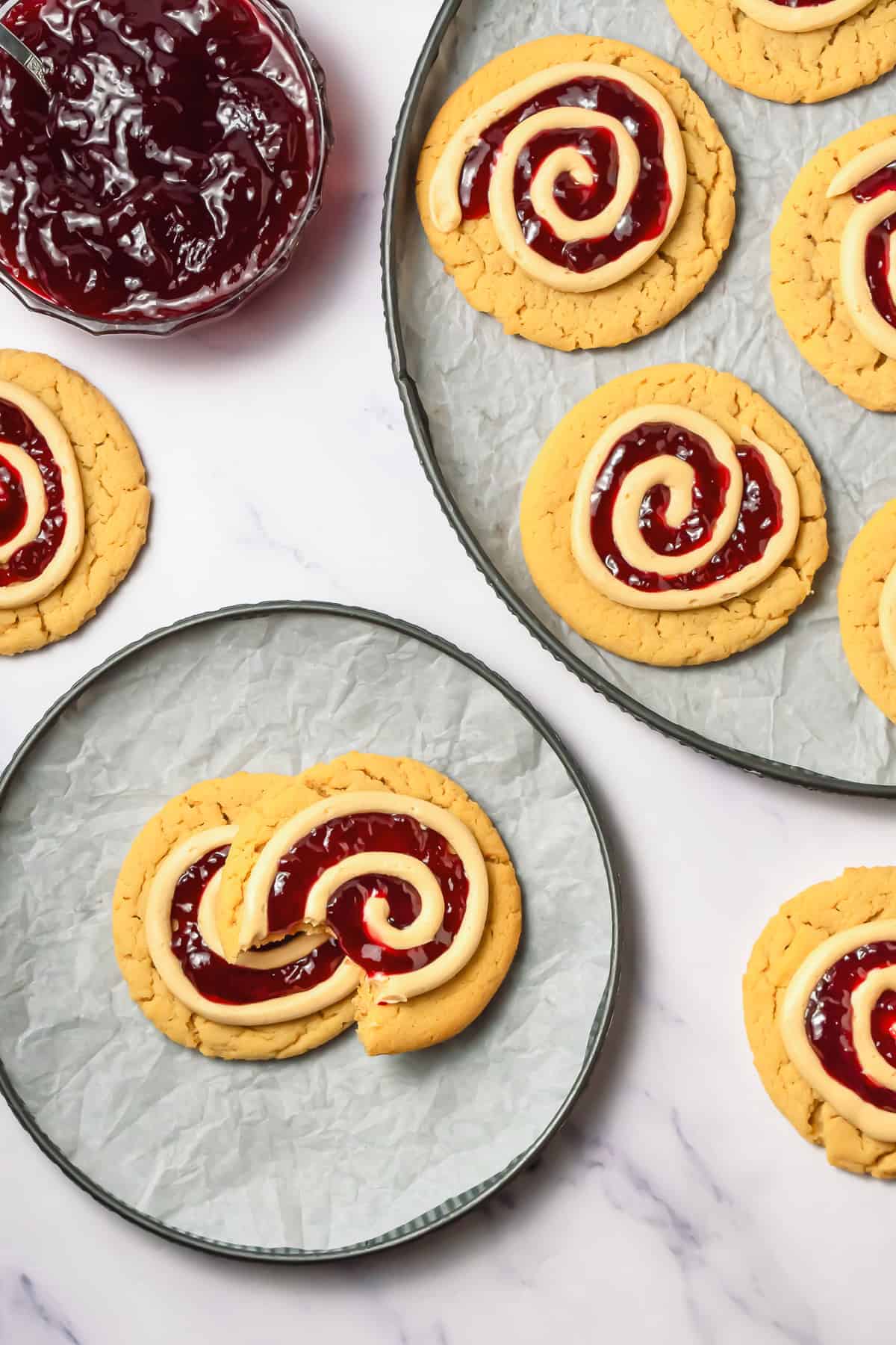 Cookies with red jelly and white icing swirls on plates, one crumbl copycat peanut butter and jelly cookie is broken, with a bowl of jelly nearby.