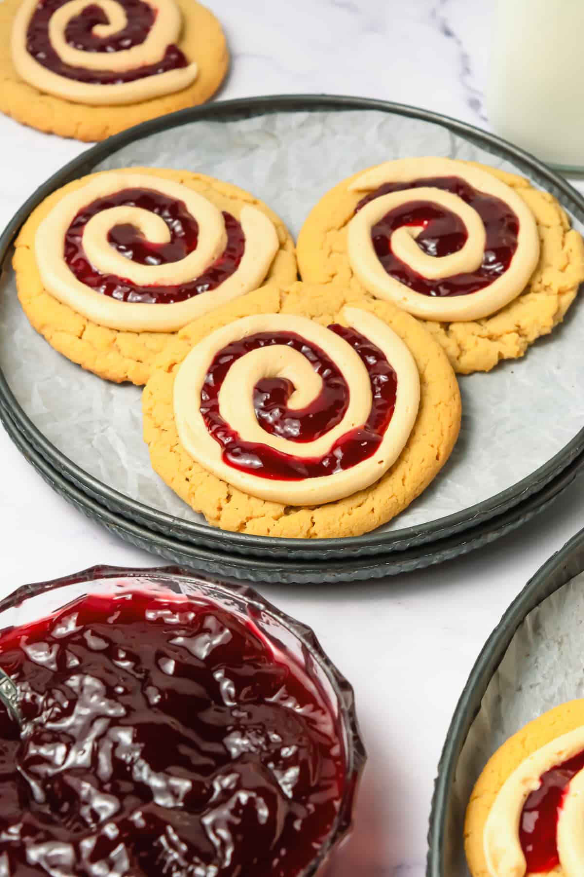 Three cookies with white icing and red jelly swirls, inspired by crumbl copycat peanut butter and jelly cookies, sit on a plate beside a jar of red jelly.