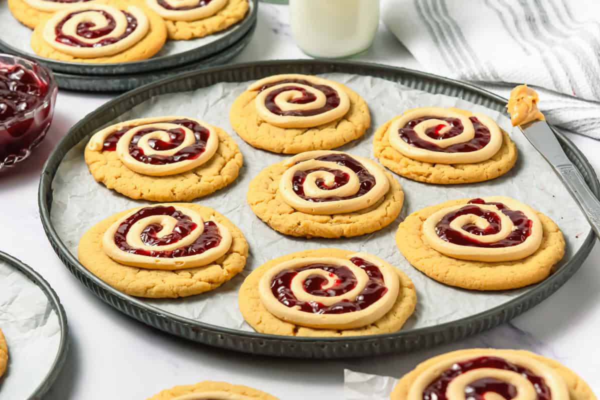 Sugar cookies topped with swirled peanut butter and jelly, inspired by Crumbl copycat peanut butter and jelly cookies, arranged on a baking tray with a glass of milk in the background.
