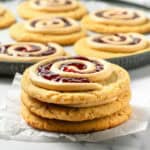 A stack of spiral sugar cookies with jelly and icing on top, inspired by crumbl copycat peanut butter and jelly cookies, with more cookies on a tray in the background.