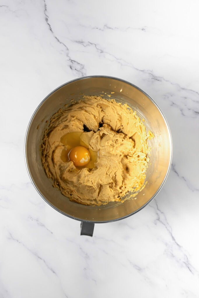 A metal mixing bowl with crumbl copycat peanut butter and jelly cookies dough and a cracked egg on top, on a marble countertop.