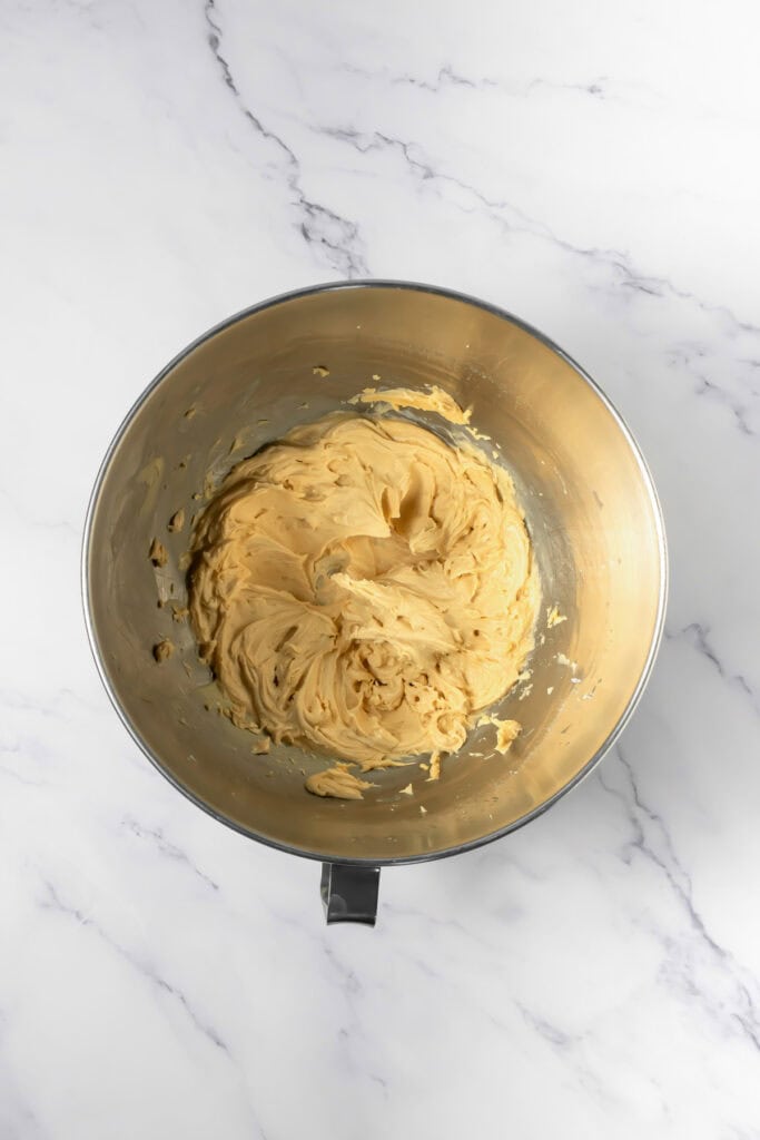 A mixing bowl filled with creamy, light brown cookie dough for crumbl copycat peanut butter and jelly cookies sits on a white marble surface.