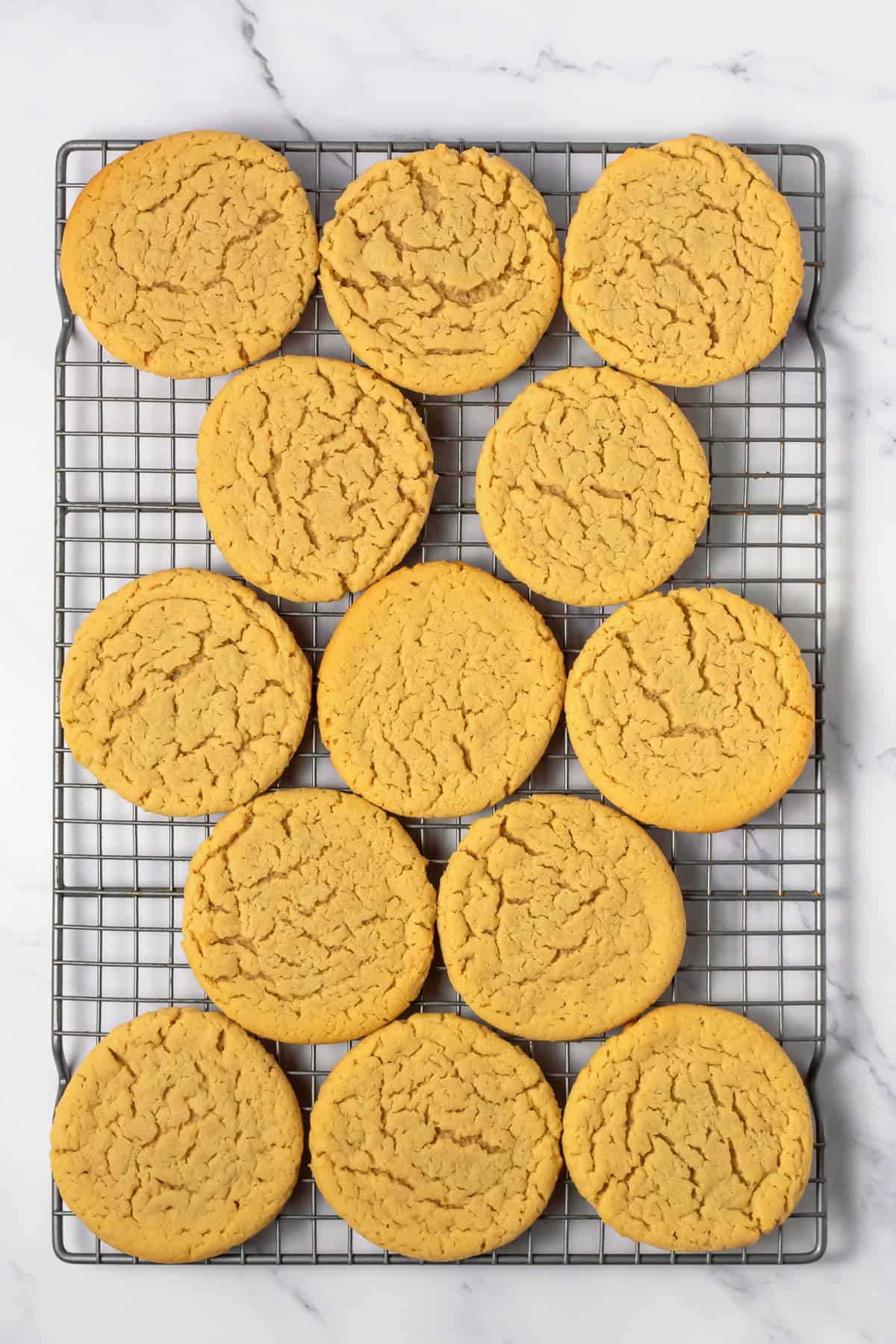 Fourteen golden brown crumbl copycat peanut butter and jelly cookies rest on a cooling rack, neatly arranged in rows on a marble surface.