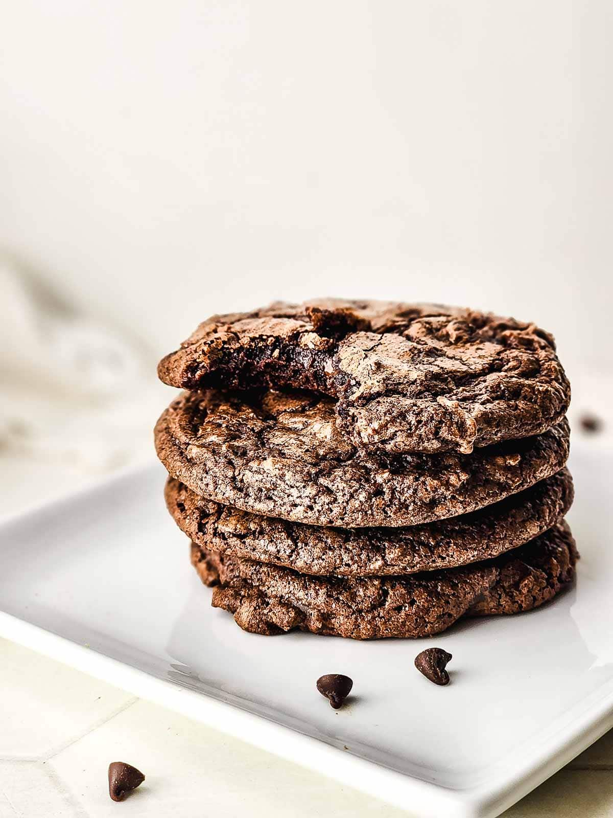 A stack of four fudgy brownie cookies sits on a white plate, with a bite taken from the top cookie.