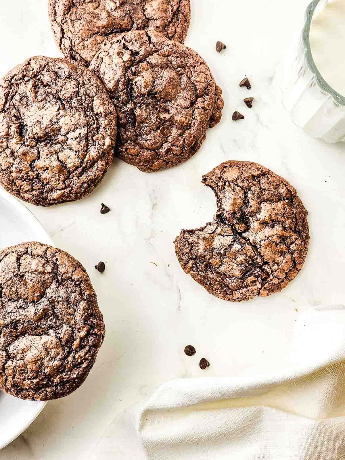 Four fudgy brownie cookies and one with a bite taken out, next to scattered chocolate chips and a glass of milk.
