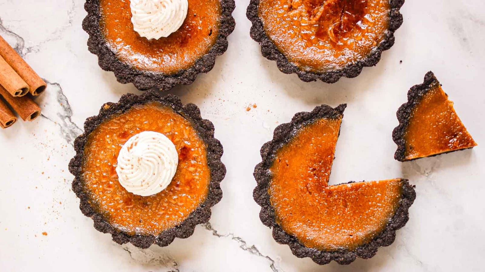 Four mini pumpkin tarts on marble, two topped with whipped cream, one with a slice missing, cinnamon sticks nearby.